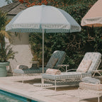 A light blue and white patterned outdoor patio umbrella shading two matching white lounge chairs with cream cushions, set beside a pool with tropical plants and a stucco wall in the background.