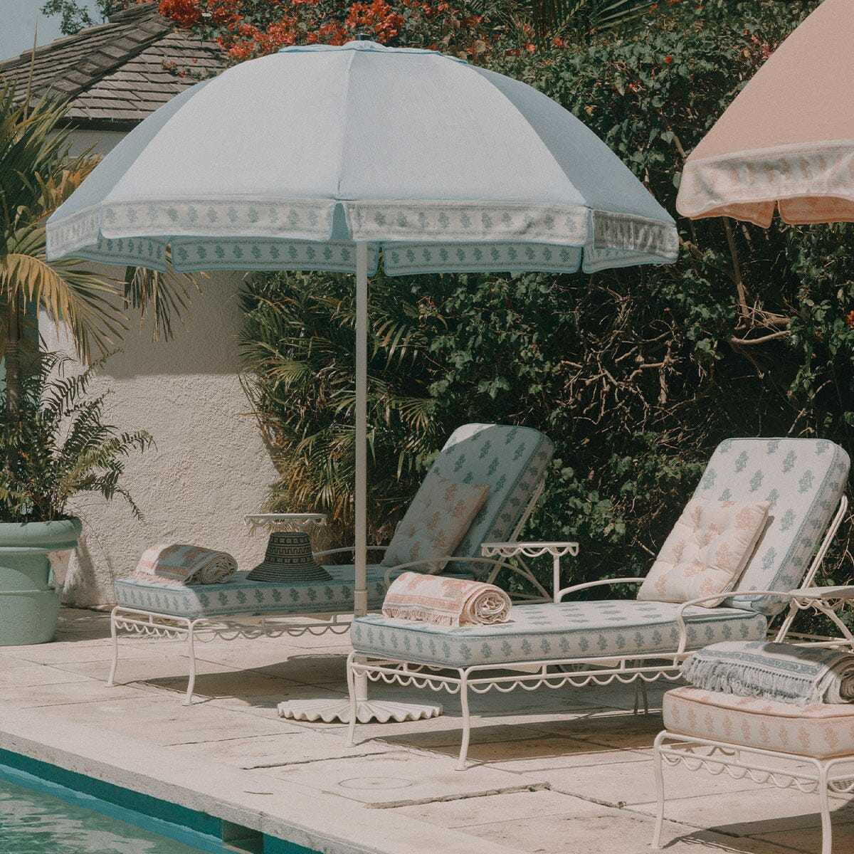 A light blue and white patterned outdoor patio umbrella shading two matching white lounge chairs with cream cushions, set beside a pool with tropical plants and a stucco wall in the background.