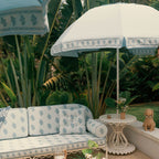 A styled outdoor patio scene featuring a white daybed and matching umbrella with a blue botanical pattern, set against a lush tropical green background.