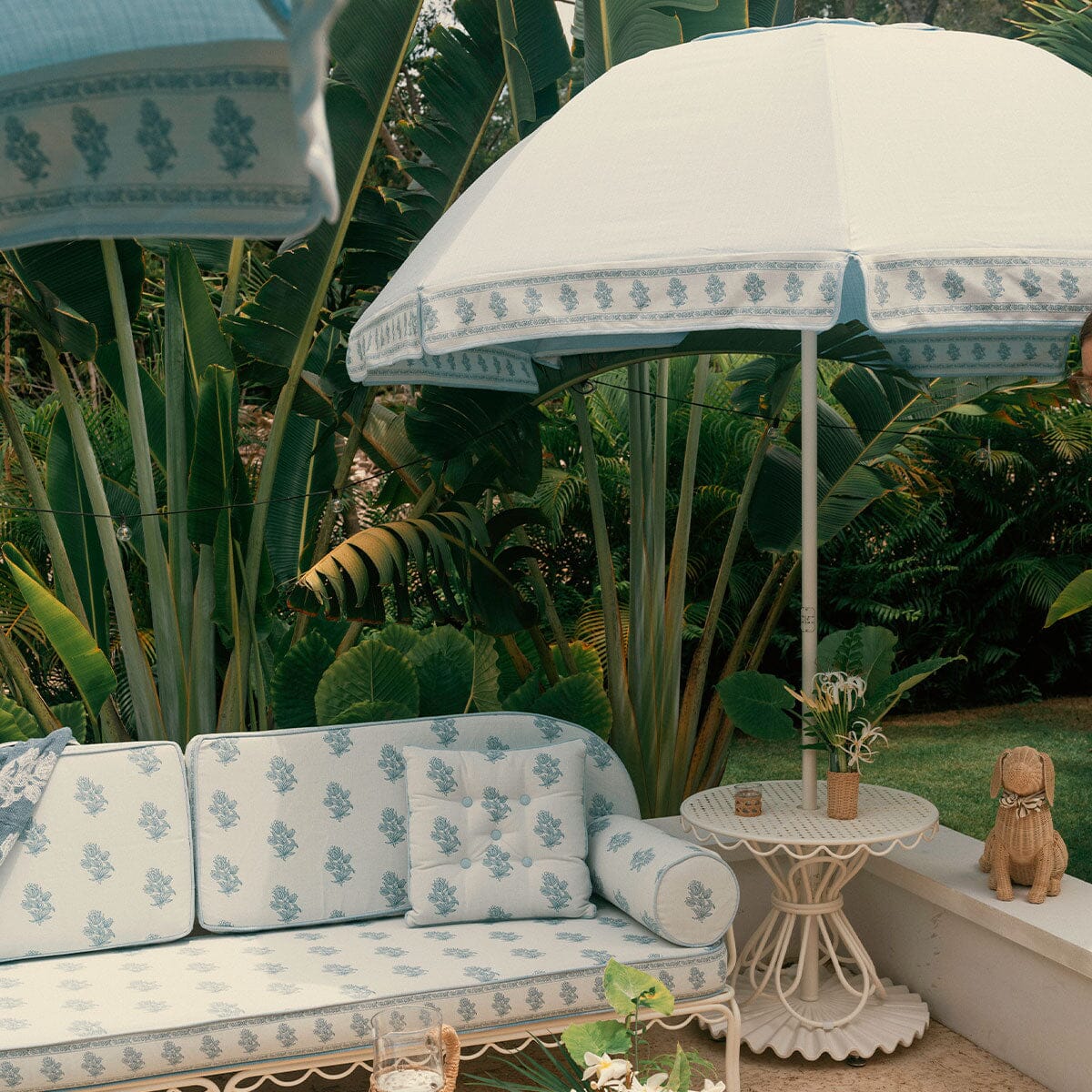 A styled outdoor patio scene featuring a white daybed and matching umbrella with a blue botanical pattern, set against a lush tropical green background.