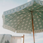 A close-up view from below of a green beach umbrella with a white floral pattern and scalloped trim, set against a pale sky with a blurred chair in the background.