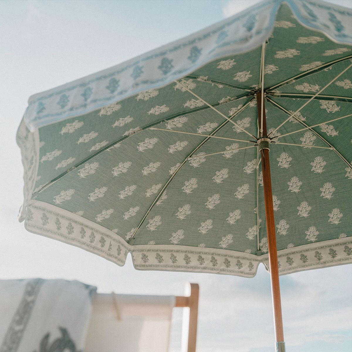 A close-up view from below of a green beach umbrella with a white floral pattern and scalloped trim, set against a pale sky with a blurred chair in the background.
