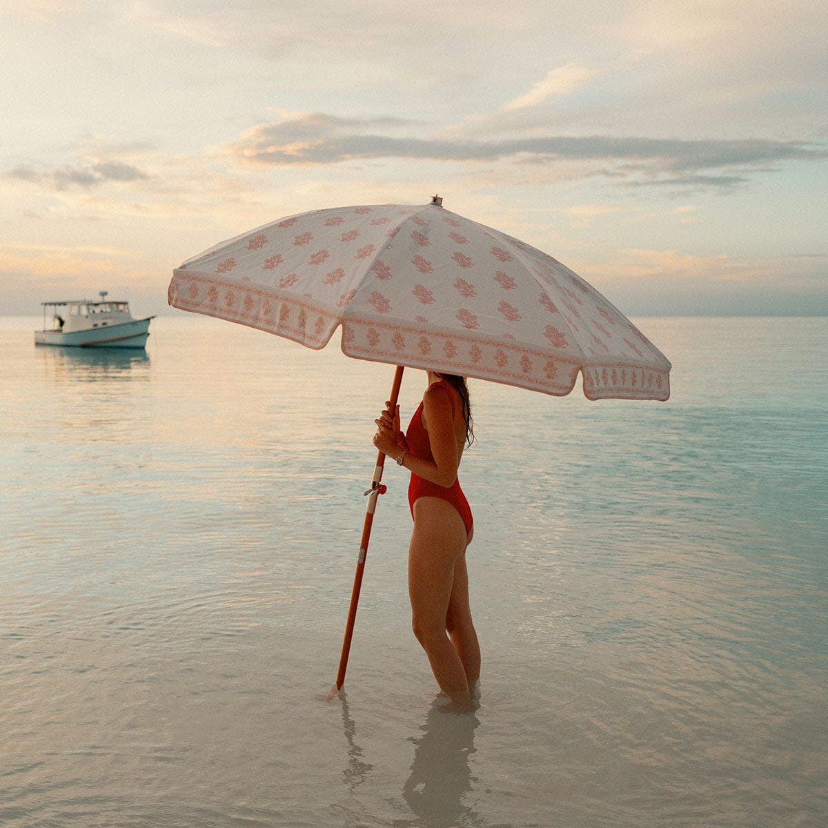 A woman in a red swimsuit stands in shallow calm water holding a large white beach umbrella with a pink floral pattern and a red handle, with a small boat and a soft sunset sky in the background.