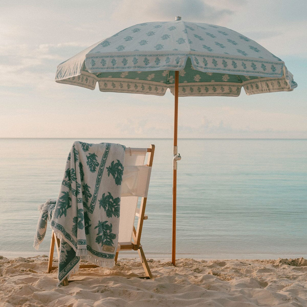 A beach scene with a large white sun umbrella featuring a green botanical pattern, a matching folded beach chair draped with a patterned towel, and pale sand under a soft blue sky.