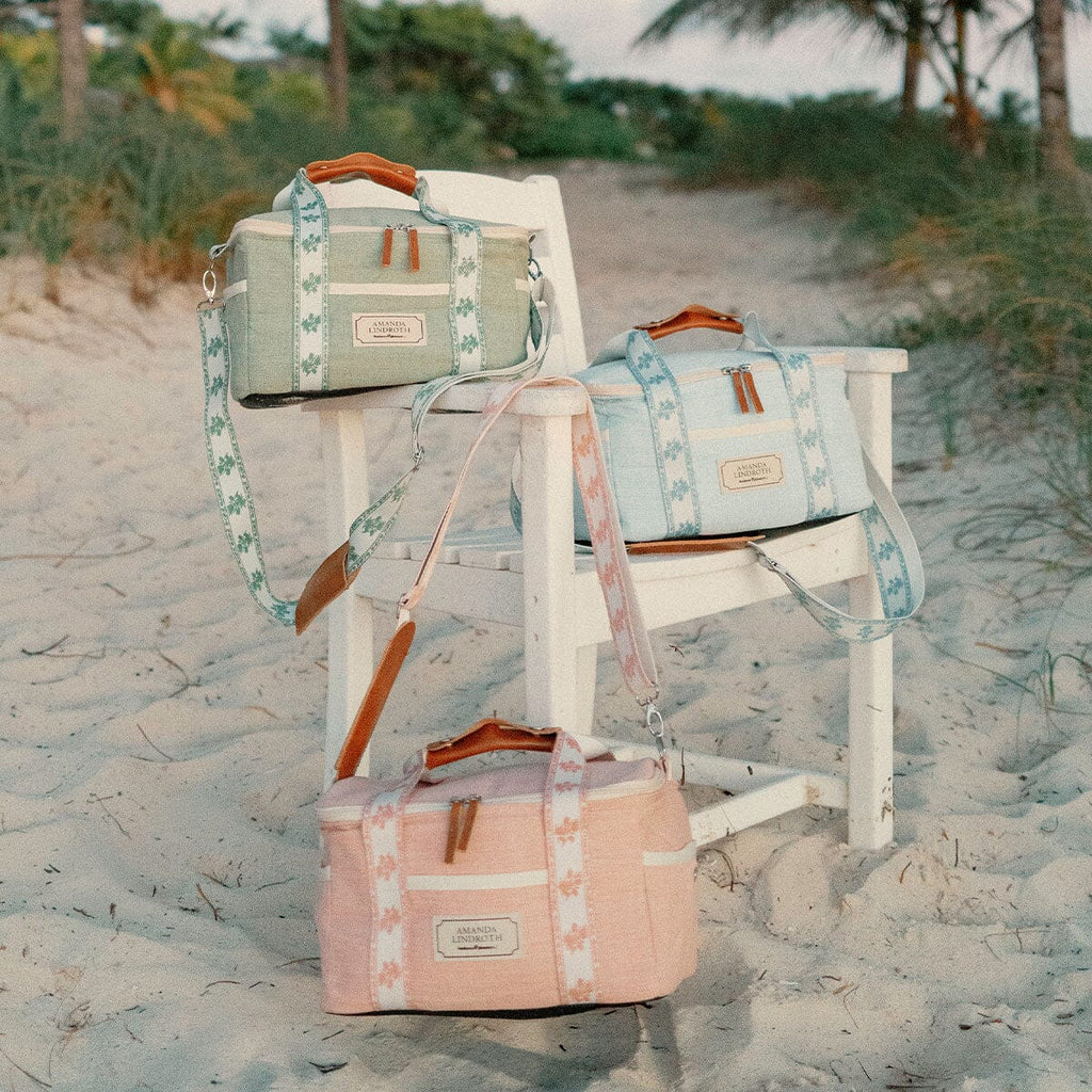 Three canvas duffel bags in pastel green, light blue, and dusty pink are arranged on a white beach chair in sandy dunes with palm trees in the background. Each bag has a floral patterned strap, brown leather handles, and a small leather brand patch.
