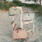 Three canvas duffel bags in pastel green, light blue, and dusty pink are arranged on a white beach chair in sandy dunes with palm trees in the background. Each bag has a floral patterned strap, brown leather handles, and a small leather brand patch.