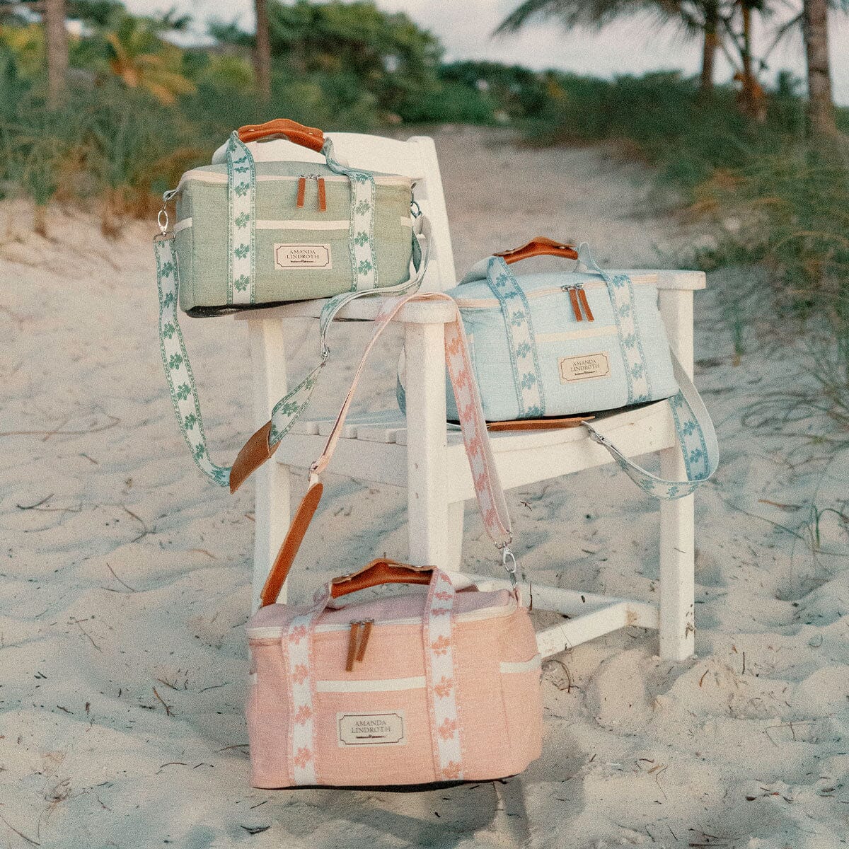 Three canvas duffel bags in pastel green, light blue, and dusty pink are arranged on a white beach chair in sandy dunes with palm trees in the background. Each bag has a floral patterned strap, brown leather handles, and a small leather brand patch.