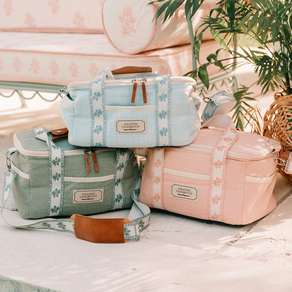 Three fabric duffel bags in pastel blue, sage green, and blush pink are stacked together on a light surface. Each bag has a floral-patterned strap, brown leather handles and zipper pulls, and a front label.