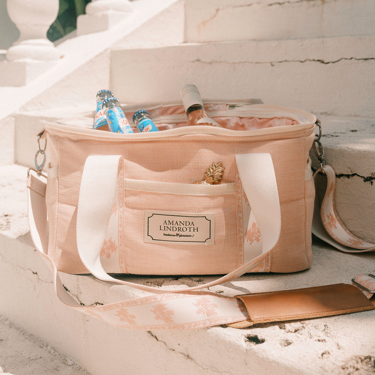 A blush pink insulated tote bag with white handles and a front pocket sits on white stone steps, filled with bottles and a drink. The front patch reads Amanda Lindroth.