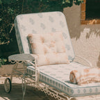 A white metal outdoor chaise lounge with light blue and beige patterned cushions, a matching pillow, and a folded throw blanket, set against a stucco wall with greenery in the background.