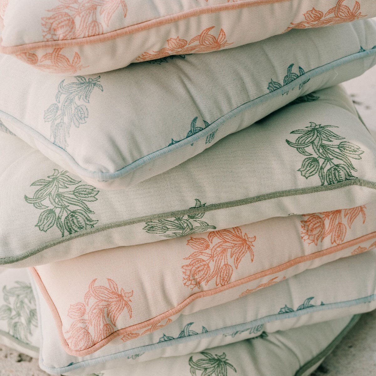 A stack of four decorative throw pillows with a botanical leaf pattern in sage green and peach, finished with light blue piping, set against a neutral stone background.