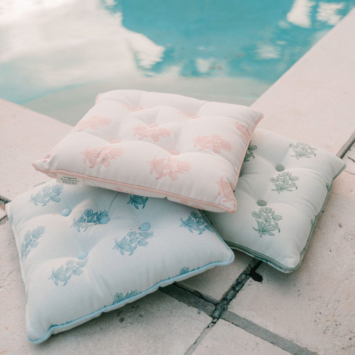 Three tufted outdoor pillows with floral prints in pastel pink, light blue, and sage green, arranged on a concrete poolside deck.