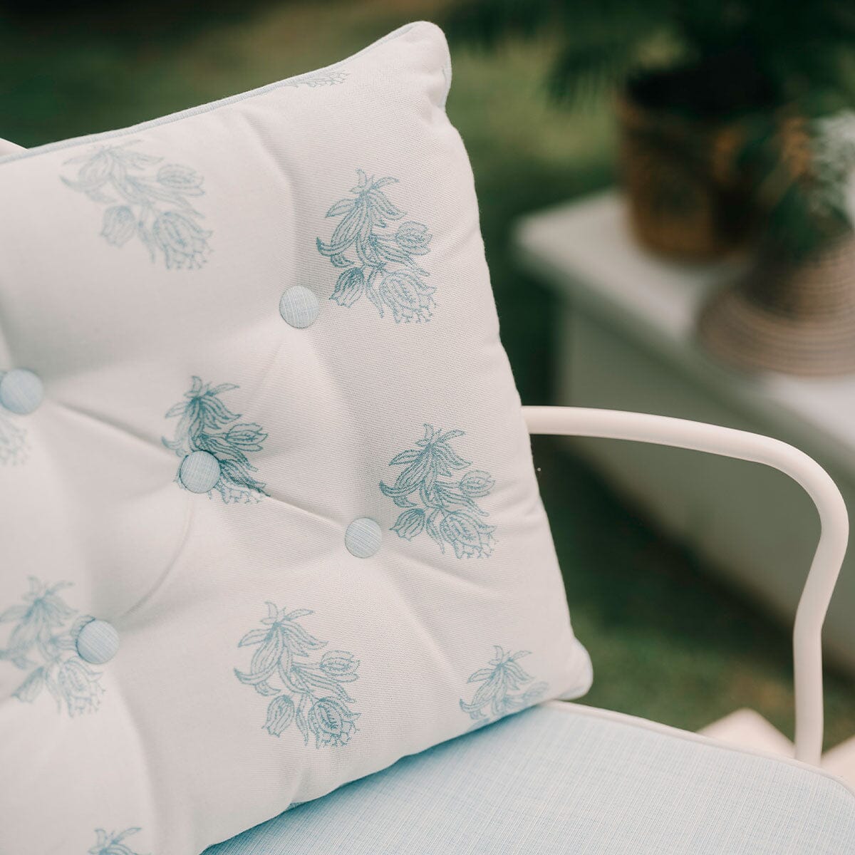 A close-up of a white tufted outdoor throw pillow with light blue floral embroidery and matching buttons, resting on a white metal chair in a garden setting.