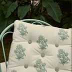 A square tufted cushion with a sage green floral pattern on an off-white background, resting on a mint green metal chair with a lush green garden backdrop.
