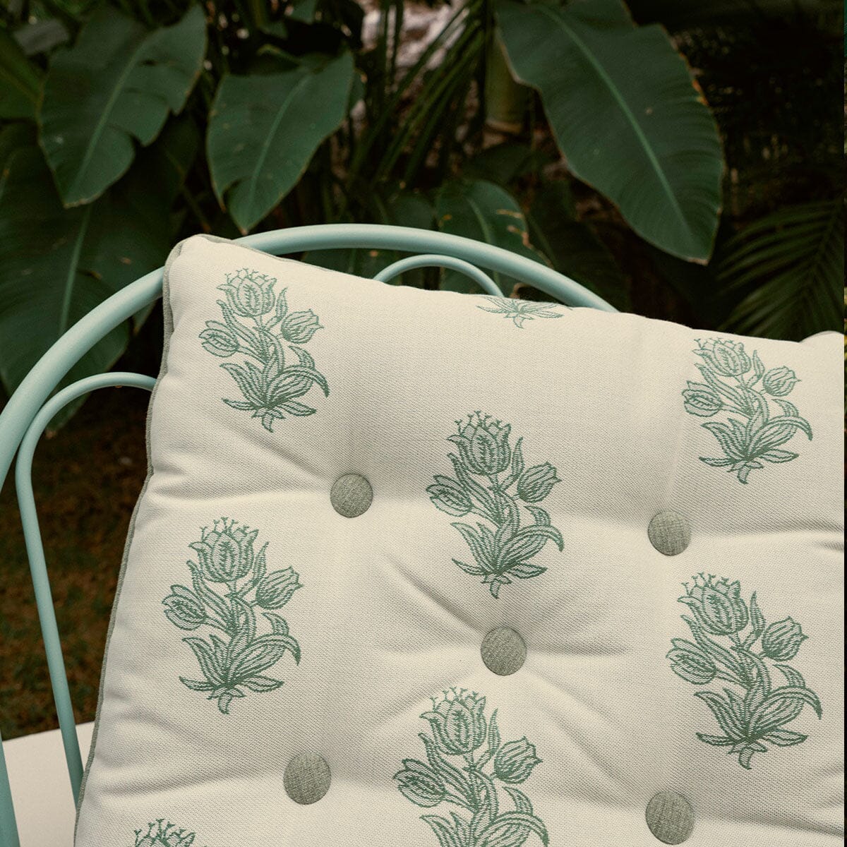 A square tufted cushion with a sage green floral pattern on an off-white background, resting on a mint green metal chair with a lush green garden backdrop.