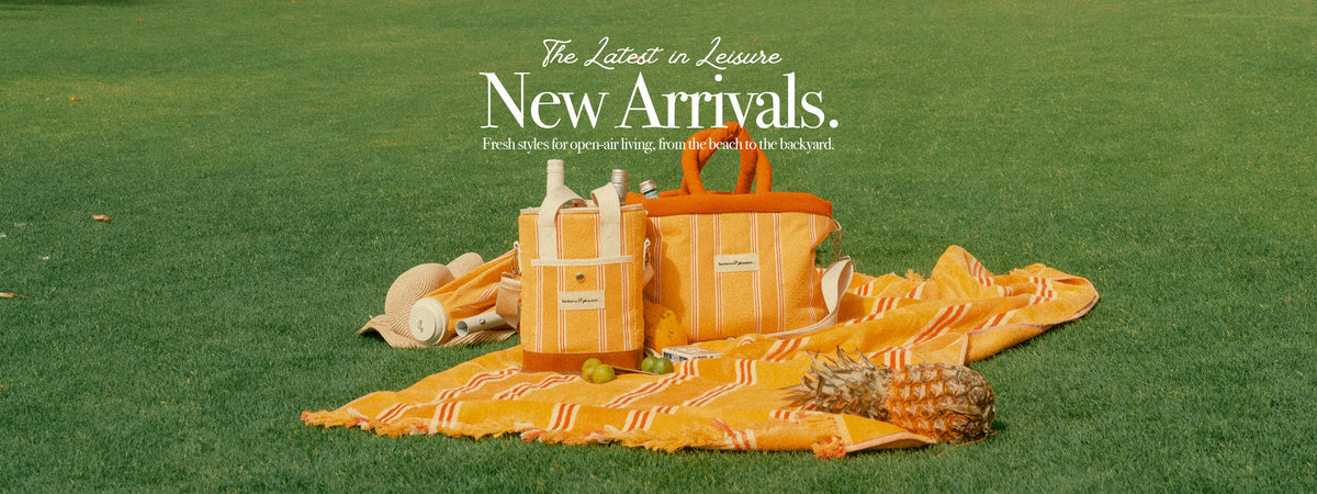A styled outdoor picnic scene on green grass featuring a yellow and orange striped blanket, matching tote bag, and matching duffel bag, with a straw hat and small accessories nearby. White promotional text at the top reads 'The Latest in Leisure New Arrivals.