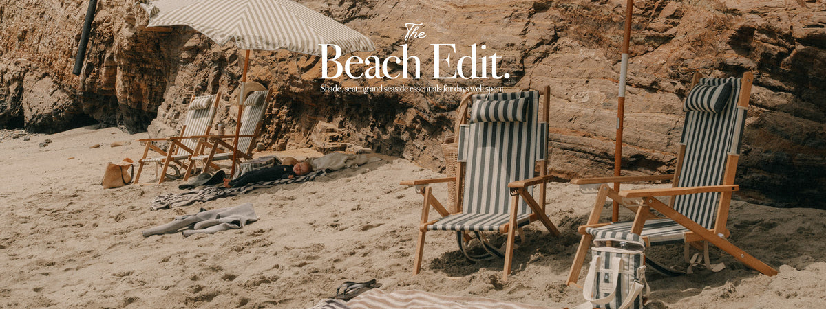 A beach scene with striped wooden deck chairs on sand in front of a rocky cliff, with an umbrella and a small table in the background. White text at the top reads 'The Beach Edit.