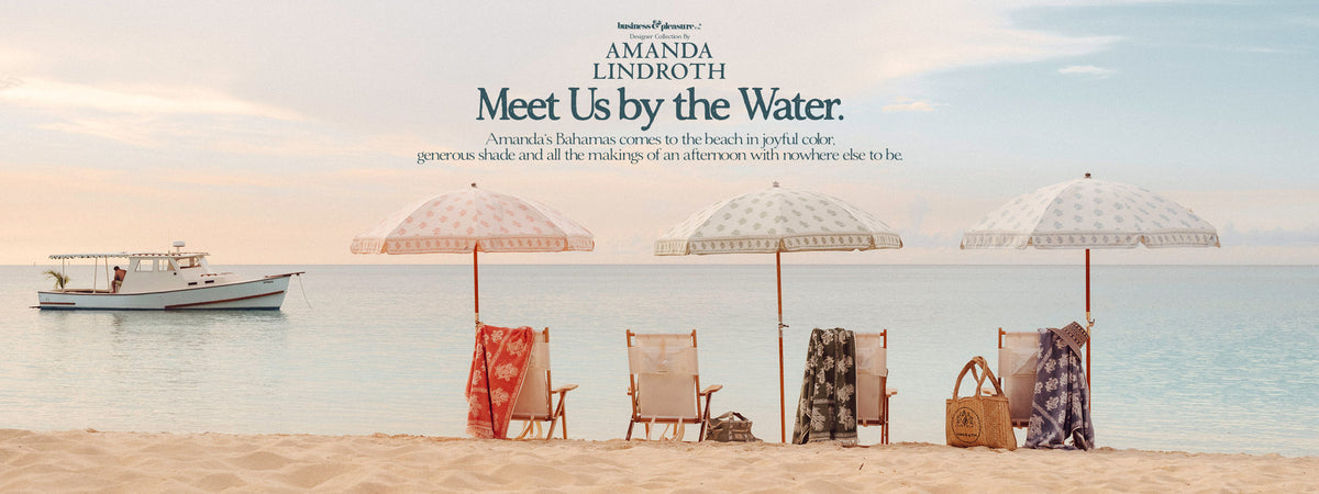A beach scene with four white umbrellas, beach chairs, towels, and a straw bag on the sand, with ocean water and a boat in the background. Text at the top reads 'AMANDA LINDROTH Meet Us by the Water.
